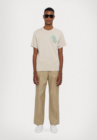 Beige cotton t-shirt with turquoise floral embroidery on the left chest, paired with beige wide-leg trousers and white sneakers. Black sunglasses.