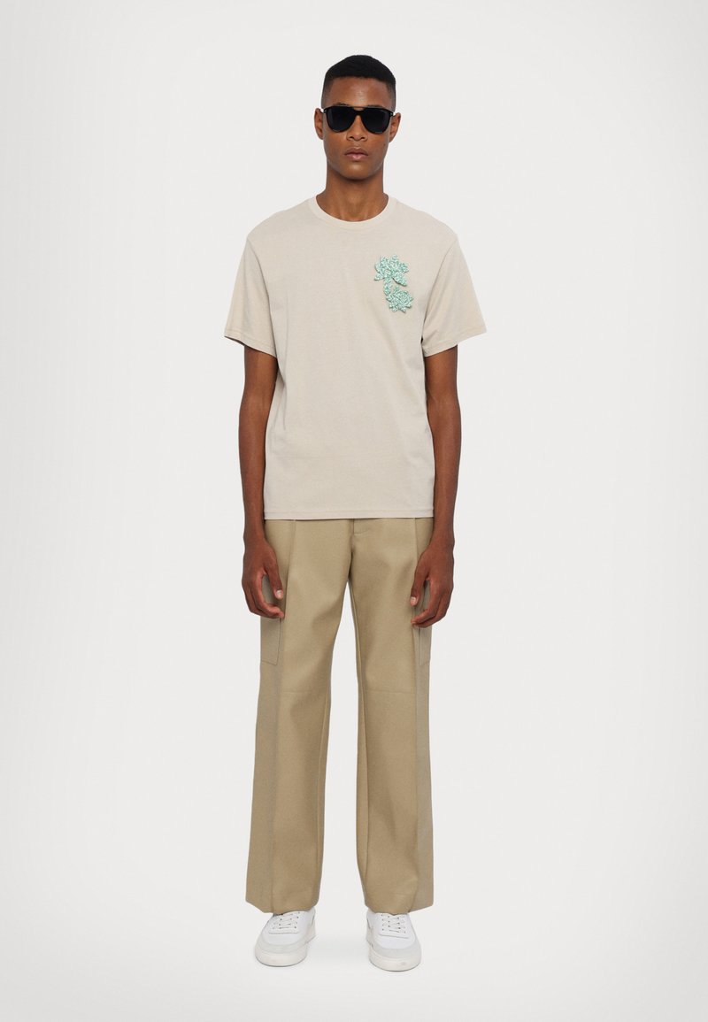 Beige cotton t-shirt with turquoise floral embroidery on the left chest, paired with beige wide-leg trousers and white sneakers. Black sunglasses.