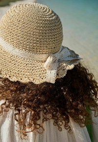 Chapeau en paille crocheté beige avec un large bord, rehaussé d'un ruban en dentelle noué à l'arrière, placé au-dessus de cheveux bruns bouclés.