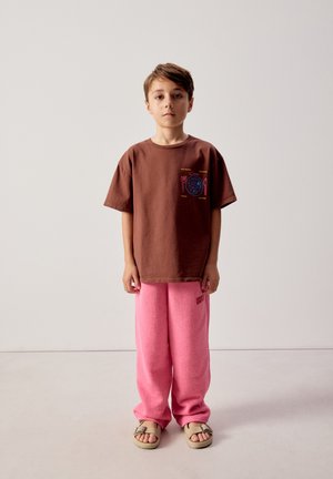 Boy standing facing forward wearing a brown t-shirt with a blue and red plate design, pink pants, and beige sandals on a plain background.