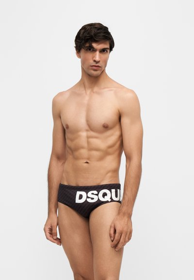 Jeune homme aux cheveux courts et foncés portant un slip de bain noir avec un lettrage blanc audacieux DSQUA, debout devant un fond uni clair.