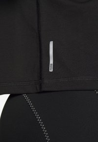 Černý sportovní top s krátkými rukávy, zdobený stříbrným logem a šitím. Měkká, pružná látka navržená pro pohodlí.