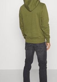Olivově zelená mikina s kapucí se stahovacími šňůrkami, žebrovanými manžetami, nošená přes tmavě šedé džíny s vybledlou texturou a dvěma zadními kapsami.