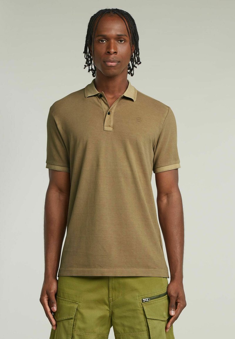 Polo shirt khaki a maniche corte, con colletto a contrasto e patta con tre bottoni. Indossato con pantaloni cargo dotati di numerose tasche.