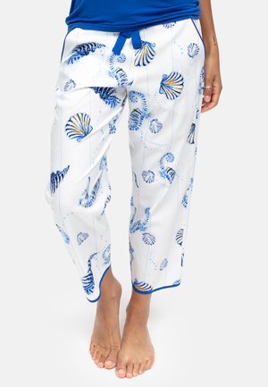 Witte pyjamabroek met blauwe en gele patronen van schelpen en zeepaardjes, blauwe bies aan de manchetten, gedragen door een staande persoon op blote voeten.
