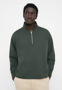 Grøn zip-up sweatshirt med høj krave, lavet af blødt materiale. Har lange ærmer og en afslappet pasform, samt syninger ved kanten.