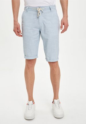 Homme portant un short bleu clair jusqu'aux genoux avec une ceinture en corde, des baskets blanches et une chemise blanche sur un fond uni.