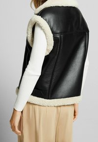 Svart imiterad läderväst med krämfärgad shearling-krage och detaljer, med en slät yta och ärmlös design. Accenterad med sid sömmar.