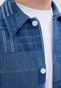 Modrá károvaná košeľa z tkaného denimového materiálu. Detailom je golier so zapínaním na gombíky a biele gombíky. Textúra je robustná s rôznymi odtieňmi modrej.