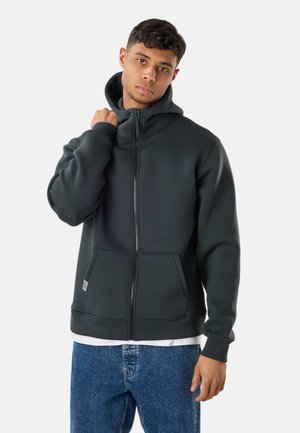 Ung mand iført en mørkegrå lynlåshoodie og blå jeans, med en hånd der justerer hoodiekraven mod en lys baggrund.