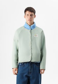 Jonge man in een lichtgroene jas met een blauwe fleece kraag en een "On Vaction" patch, gecombineerd met blauwe jeans tegen een witte achtergrond.