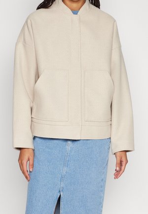 Personne portant une veste à manches longues couleur crème avec poches à l'avant et une jupe en jean bleu clair avec une fente devant.