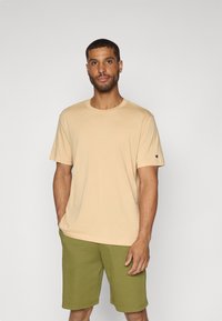 Beige bomull T-skjorte med korte ermer og rund hals, kombinert med grønne shorts. T-skjorten har en liten logo på venstre erme.