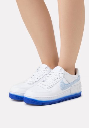 Valkoiset nauhalliset tennarit, joiden pohjissa on sininen liukuvärjäys ja tekstuurilliset vaaleansiniset Swoosh-logot, jalassa ilman sukkia valkoista taustaa vasten.