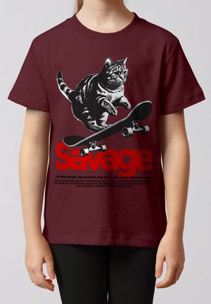 Een gestreepte kat die op een skateboard springt boven het opvallende rode woord "Savage" op een bordeauxrood T-shirt gedragen door een kind dat naar voren kijkt.