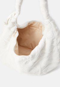 Hvit strukturt veske med myk stoffoverflate, glidelås lukking og beige innvendig fôr. Håndtakene har et vridd design.