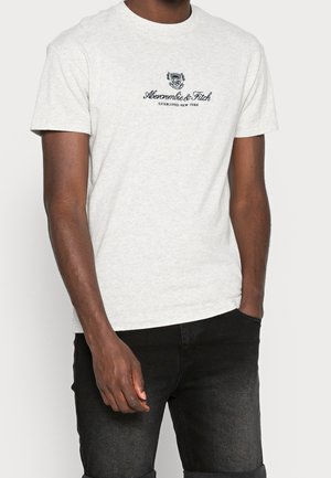 T-shirt en coton gris clair avec un col rond, des manches courtes et un imprimé logo sur le devant en bleu marine. À associer avec un pantalon en denim foncé.