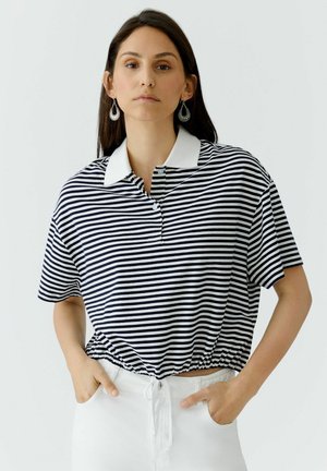 Jonge vrouw met lang donker haar, gekleed in een zwart-wit gestreept polohemd met korte mouwen en witte broek, poserend met haar handen in haar zakken.