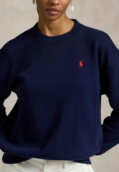 Γυναίκα που φοράει μπλε ναυτικού χρώματος πουλόβερ με λαιμόκοψη και κόκκινο λογότυπο Polo Ralph Lauren, καθώς και σκουλαρίκια-κοχύλια, συνδυασμένο με λευκό παντελόνι.