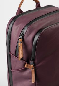 Zaino marrone con cerniere nere, dettagli in pelle e una forma strutturata. Presenta scomparti con doppia zip e una tasca laterale in rete.