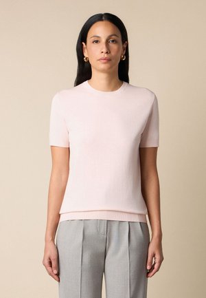 Kobieta z długimi ciemnymi włosami, nosząca złote kolczyki koła, blado różowy sweter z krótkim rękawem oraz jasnoszare plisowane spodnie, stojąca na beżowym tle.