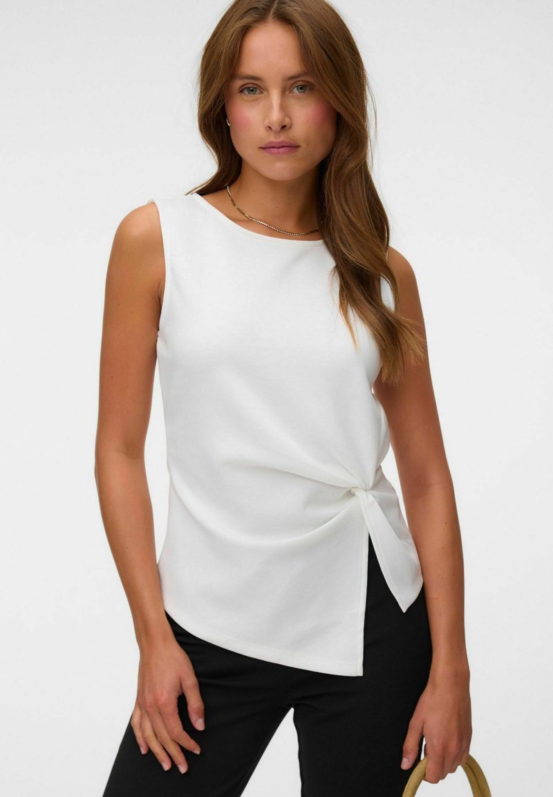 Femme aux longs cheveux bruns portant un haut blanc sans manches avec un nœud sur le côté et un pantalon noir, tenant un sac à main beige sur fond blanc.