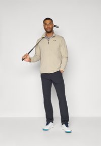 Veste zippée beige sur un polo gris, associée à un pantalon sombre et des chaussures de golf blanches. Le mannequin tient un club de golf, se tenant devant un fond uni.