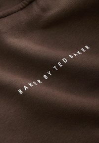 Hnědá bavlněná látka s textem "BAKER BY TED BAKER" v bílé barvě, umístěným horizontálně přes materiál. Měkká textura, jemný lesk.