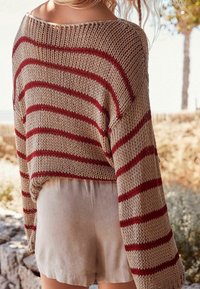 Femme portant un pull beige ample en maille avec des rayures rouges et un short beige clair à l'extérieur, près d'arbres et d'un mur en pierre.