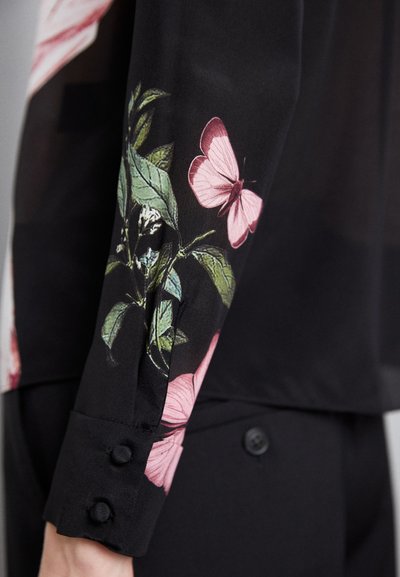 Blusa nera con motivo floreale, con farfalle rosa e foglie verdi sulle maniche, arricchita da dettagli con bottoni neri.