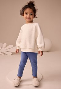 Bluză albă cu pete, cu mâneci largi, colanți albaștri cu pete în formă de nori și papuci pufosi de culoare crem.