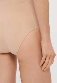 Nude beškoniai aukšti apatiniai, su glotnia tekstūra, elastingu diržu ir nematomis siūlėmis. Siūlomas vidutinio aukščio kirpimas ir minimalistinis dizainas.