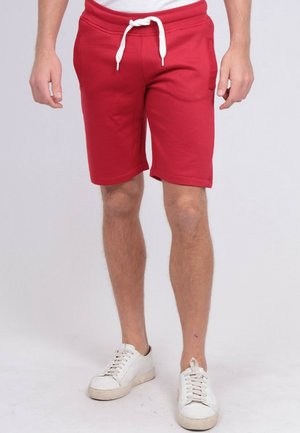 Homme portant un short rouge avec cordon et des baskets blanches, debout devant un fond uni.