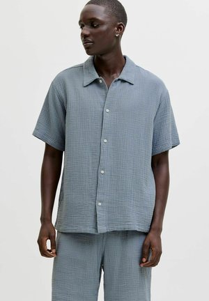 Osoba o ciemnej karnacji ubrana w luźną, jasnoniebieską, teksturowaną koszulę na krótki rękaw zapinaną na guziki oraz pasujące szorty, spoglądająca na bok na tle gładkiego tła.