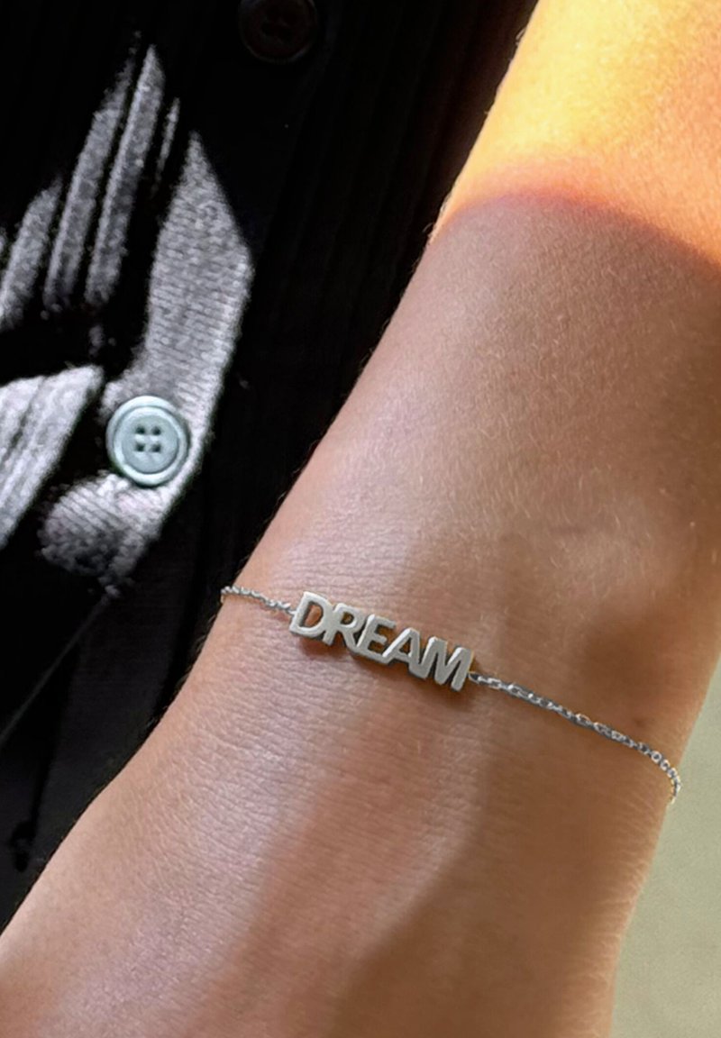 Sidabro spalvos grandinėlės apyrankė su žodžiu „DREAM“ didžiosiomis raidėmis, dėvima ant šviesios odos tono riešo. Paprastas dizainas, subtili tekstūra.