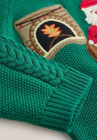 Grüner gestrickter Pulloverärmel mit geflochtenem Muster, geripptem Bündchen und gesticktem Patch, der einen Ziegelbogen mit einem Feuersymbol und einem Teil einer roten Figur zeigt.