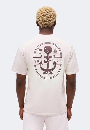 Person mit kurzen lockigen blonden Haaren trägt ein hellbeiges T-Shirt mit großem Anker-, Mond-, Flaggen- und Segelboot-Grafik mit "2026" auf dem Rücken.
