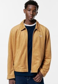 Giacca zip-up in suede color sabbia con colletto, tasche laterali e dettaglio ricamato "Nyc" sul petto, indossata sopra una maglietta a girocollo blu navy.