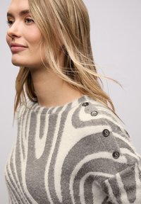 Pull gris et blanc à motif, fabriqué en maille douce, avec un col rond et des boutons décoratifs foncés sur une épaule.