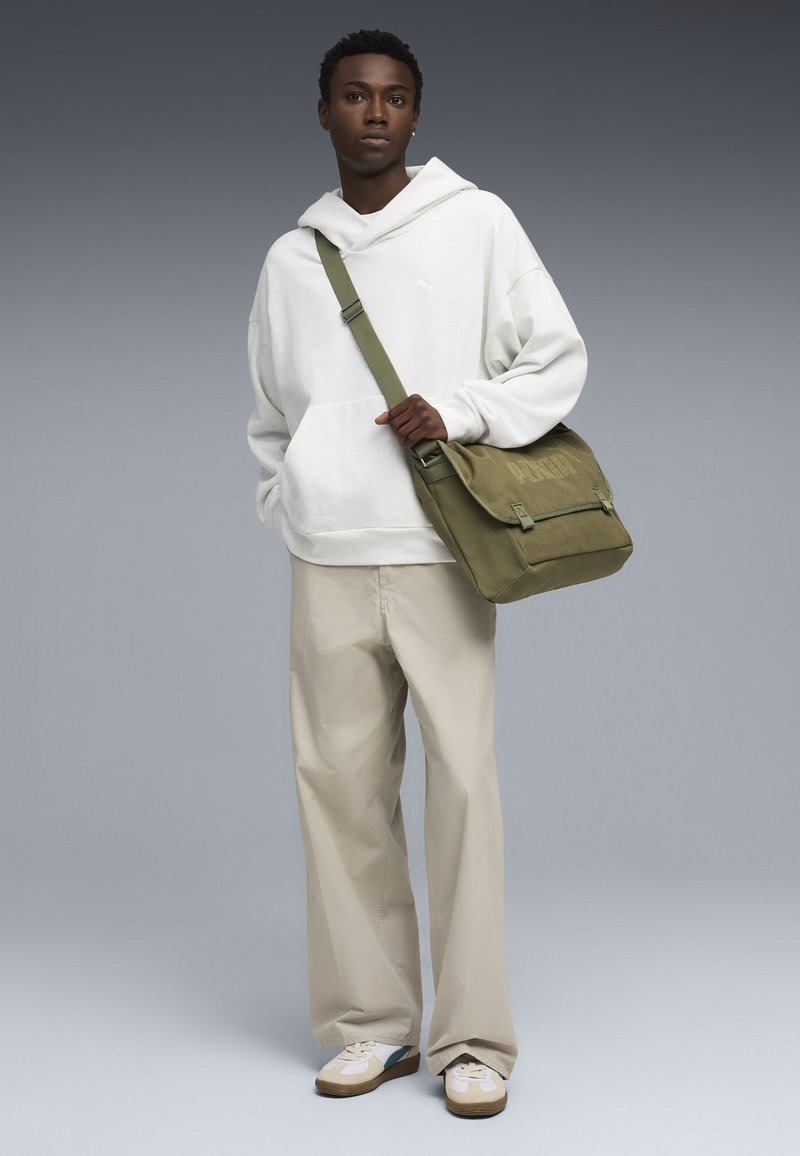 Jeune homme portant un sweat à capuche blanc, un pantalon beige, des baskets blanches et un sac à bandoulière vert olive sur un fond gris.