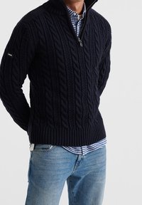 Marinblå kabelstickad tröja med hög krage och dragkedja halvvägs, ribbat nederkant, bärs över en blå och vit rutig skjorta, tillsammans med ljusa jeans.