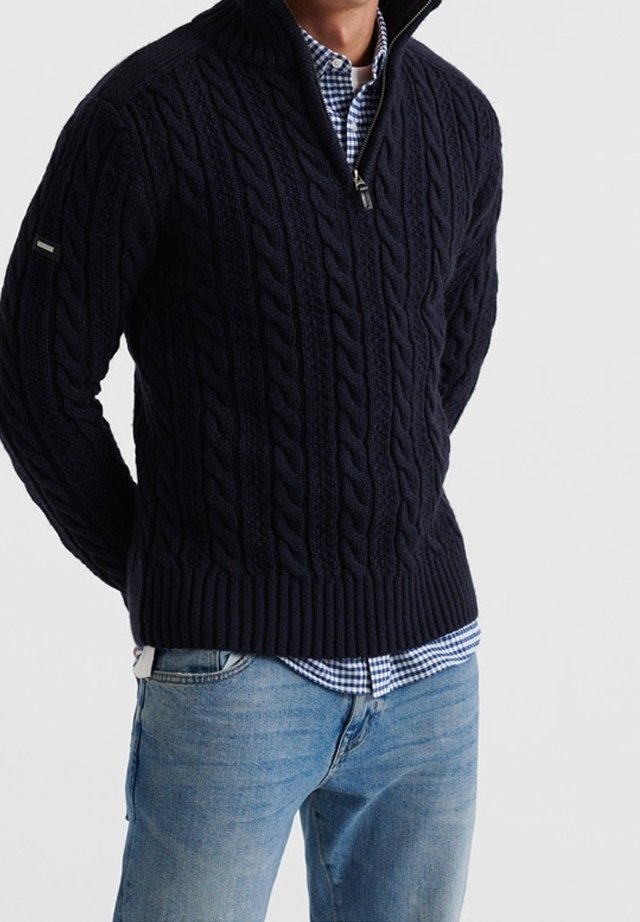 Marinblå kabelstickad tröja med hög krage och dragkedja halvvägs, ribbat nederkant, bärs över en blå och vit rutig skjorta, tillsammans med ljusa jeans.