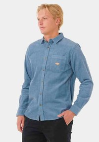 Camisa de pana azul claro con diseño de botones delanteros, bolsillo en el pecho y parche de logo marrón. Mangas largas y tejido texturizado.