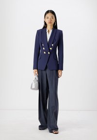 Marineblaues, doppelt geknöpftes Blazer-Jackett mit goldenen Knöpfen, kombiniert mit maßgeschneiderten grauen Hosen. Das Model hält eine kleine silberne Handtasche und trägt große Creolen.