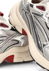 Zapatillas deportivas plateadas y crema con material de malla, suelas de goma y acentos rojos. Presentan un diseño texturizado y una marcada marca en el lateral.
