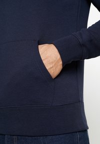 Hand delvis inskjuten i framfickan på en marinblå sweatshirt, bärs tillsammans med blå jeans.