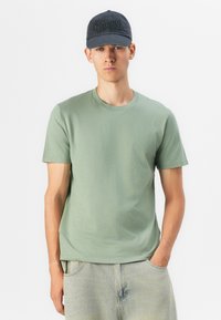 T-shirt din bumbac verde deschis cu guler rotund și mâneci scurte, asortat cu blugi de denim deschis. Modelul poartă o șapcă închisă, cu litere texturate.