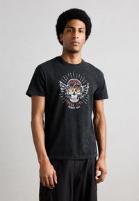 Homme avec une coiffure afro portant un T-shirt noir avec un graphisme de crâne en flammes et des ailes, ainsi qu'un pantalon cargo noir, debout devant un fond uni.