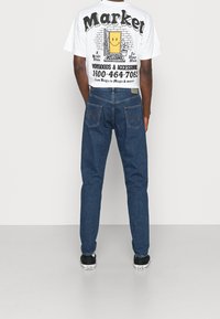 T-shirt bianco con grafica che presenta una bevanda in stile cartoon con testo sulla schiena; abbinato a jeans in denim blu e scarpe nere con suole bianche.