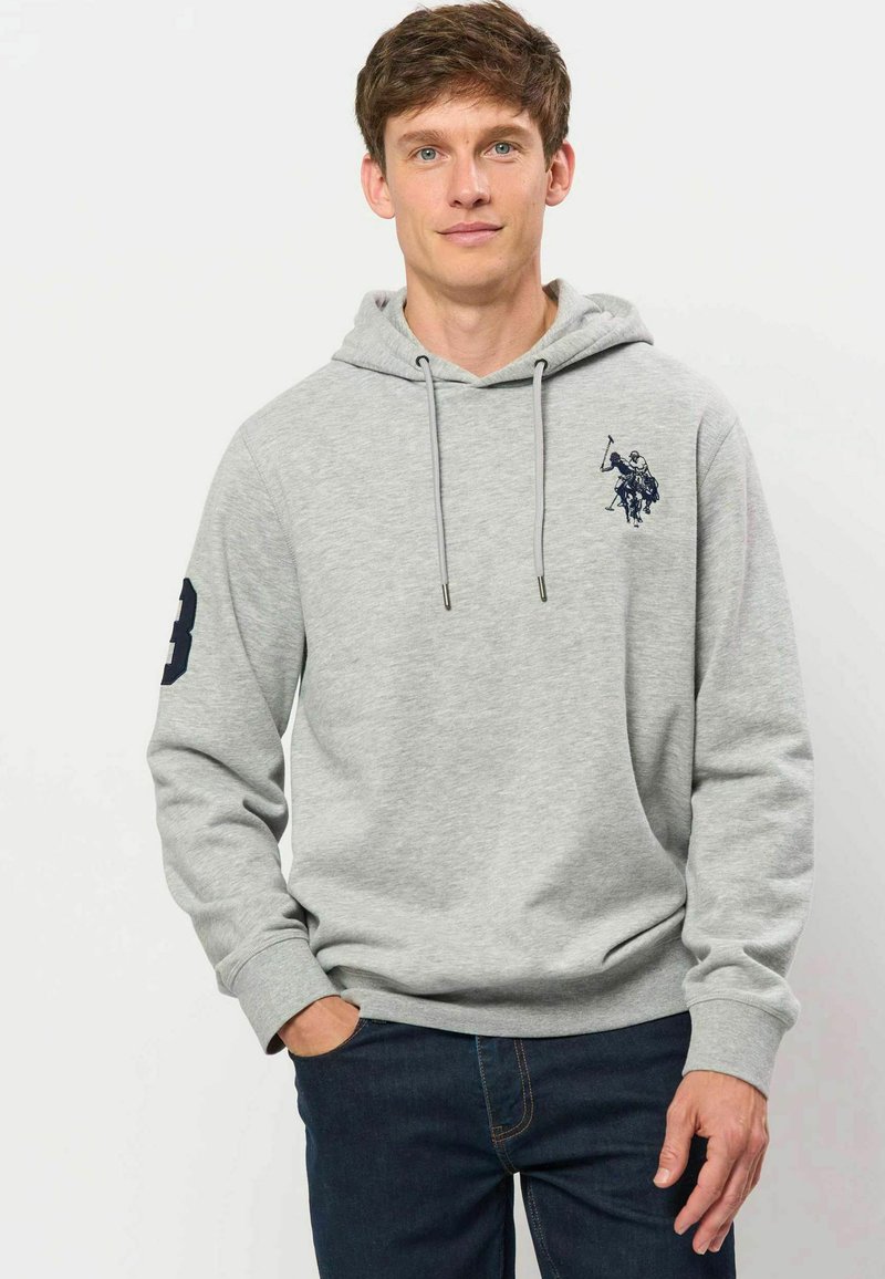 Homme portant un sweat à capuche gris clair avec un logo de joueur de polo sur la poitrine et le numéro huit sur la manche gauche.